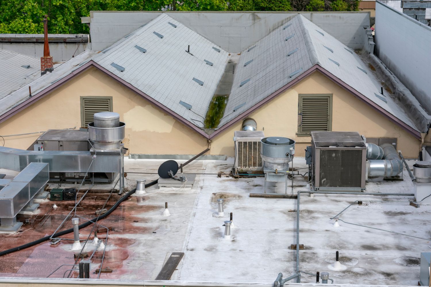 A rooftop with various HVAC units, vents, and a satellite dish, surrounded by other buildings. The surface shows signs of wear and discoloration, potentially in need of roof replacements & repairs. A rooftop with various HVAC units, vents, and a satellite dish, surrounded by other buildings The surface shows signs of wear and discoloration, potentially in need of roof replacements & repairs