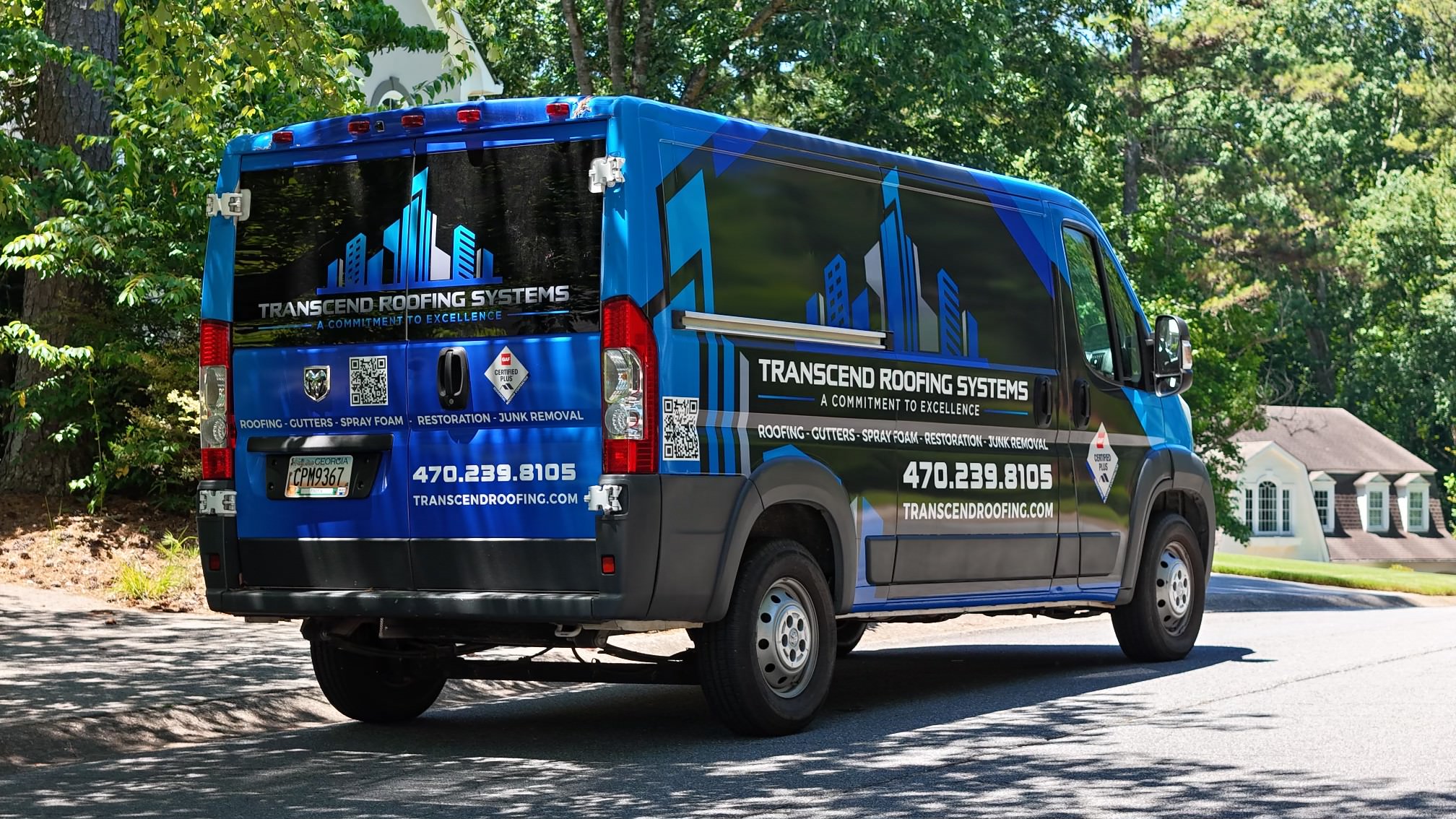 A blue Transcend Roofing Systems van is parked on a residential street, showcasing detailed company graphics and contact information on its sides and back, proudly advertising Roof Replacements & Repairs. A blue Transcend Roofing Systems van is parked on a residential street, showcasing detailed company graphics and contact information on its sides and back, proudly advertising Roof Replacements & Repairs