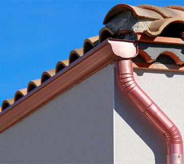 Terracotta roof tiles on a house with a pink metal rain gutter system, standing out against a clear blue sky—ideal for seamless roof replacements and repairs.