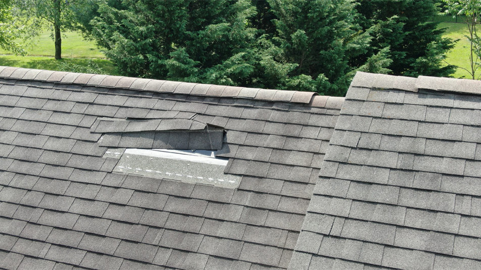 Damaged asphalt shingle roof with missing and displaced shingles exposing the underlayment, surrounded by trees in the background 