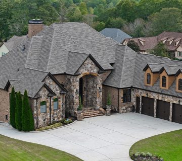 Large stone and brick house with a three car garage, steep roof, and driveway, surrounded by grass and neighboring homes in a suburban area 