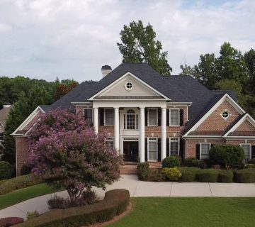 A large, two story brick house with white columns, black shutters, and manicured landscaping, surrounded by trees and a curved driveway 