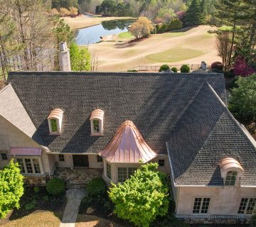 Aerial view of a large house with a dark shingled roof, surrounded by trees, and a golf course with a pond in the background 