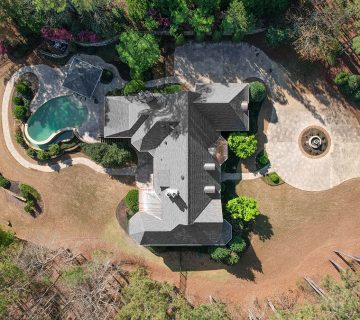 Aerial view of a large house with a driveway, circular fountain, landscaped yard, trees, and a backyard pool 