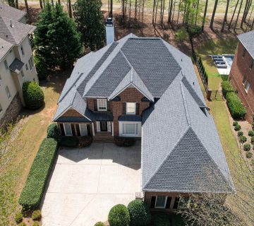 Aerial view of a two story brick house with a grey shingled roof, double garage, and a driveway, surrounded by neighboring homes and trees 