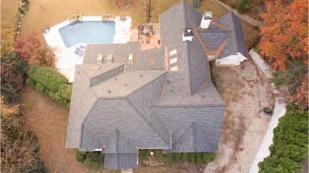 Aerial view of a house with a grey shingle roof, surrounding patio, pool, driveway, and autumn trees.