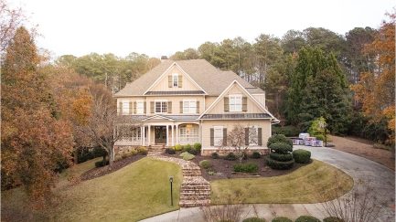 Two-story beige house with a large front lawn, stone walkway, and surrounding trees, seen from above on an overcast day.
