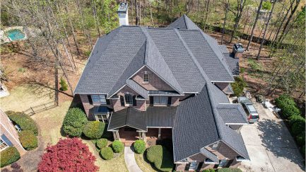 Aerial view of a two-story brick house with a black shingle roof, front porch, driveway, and surrounding trees and shrubs.