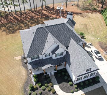 Aerial view of a large two story house with gray roof, white exterior, driveway, parked cars, and surrounding lawn with trees