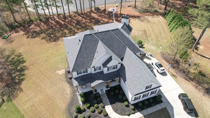 Aerial view of a large two-story house with gray roof, white exterior, driveway, parked cars, and surrounding lawn with trees.