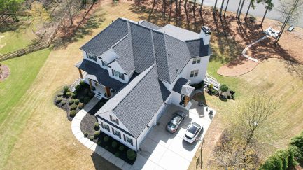 Aerial view of a large two-story house with dark roof shingles, two cars parked in the driveway, and landscaped yard surrounded by trees and grass.