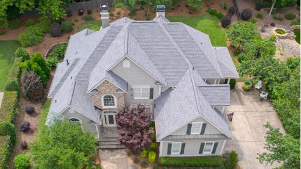 Aerial view of a large two-story gray house with multiple gabled roofs, surrounded by landscaped greenery and a driveway.