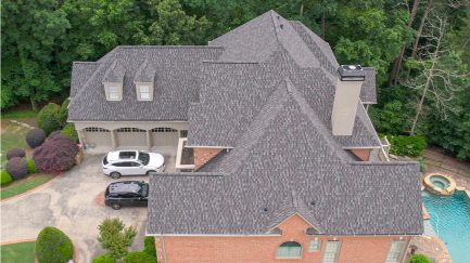 Aerial view of a large brick house with gray shingle roof, three-car garage, two parked cars, swimming pool, and surrounding trees.