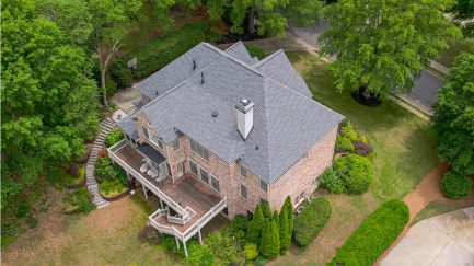 Aerial view of a two-story brick house with a large wooden deck, surrounded by trees and landscaped greenery.