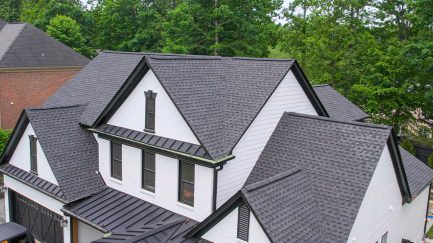 Aerial view of a house with multiple gabled roofs covered in gray asphalt shingles, featuring white siding and black trim, surrounded by trees.