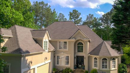 Two-story beige house with multiple gables, arched windows, and a dark shingle roof, surrounded by trees under a partly cloudy sky.