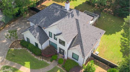 Aerial view of a two-story house with gray shingles, white exterior walls, a curved driveway, and landscaped yard with bushes and trees.