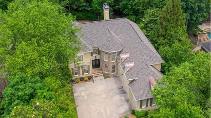 Aerial view of a large, two-story house with a stone and stucco exterior, surrounded by dense trees, and a spacious concrete driveway in front.