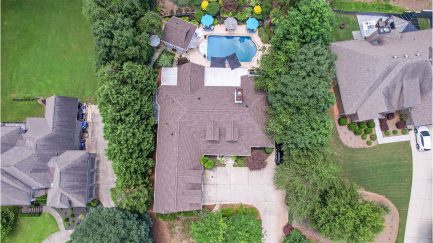 Aerial view of a suburban home with a backyard pool, patio, and surrounding trees, situated between neighboring houses with lawns and driveways.