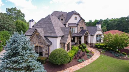 A large two-story stone and brick house with multiple peaked roofs, surrounded by landscaped bushes, trees, and a curved walkway.