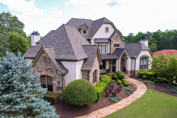 A large two-story stone and brick house with multiple peaked roofs, surrounded by landscaped bushes, trees, and a curved walkway. A large two-story stone and brick house with multiple peaked roofs, surrounded by landscaped bushes, trees, and a curved walkway.