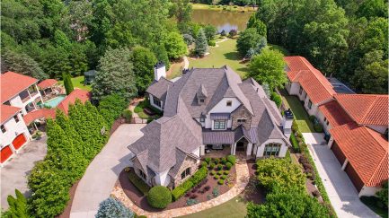 Aerial view of a large, gray-roofed house with landscaped yard, circular driveway, and nearby buildings, surrounded by trees and a lake in the background.