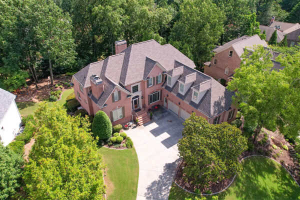 Aerial view of a large two-story brick house with a gray roof, surrounded by trees, landscaped yard, and a curved driveway in a suburban neighborhood. Aerial view of a large two-story brick house with a gray roof, surrounded by trees, landscaped yard, and a curved driveway in a suburban neighborhood.