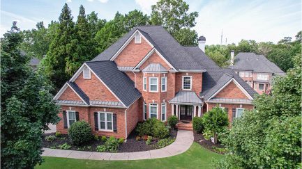Two-story brick house with multiple gables, black shingle roof, white trim, landscaped front yard, and surrounding trees, photographed from above.