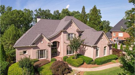Two-story brick house with multiple gables, large front windows, manicured bushes, and a curving sidewalk, surrounded by trees on a sunny day.