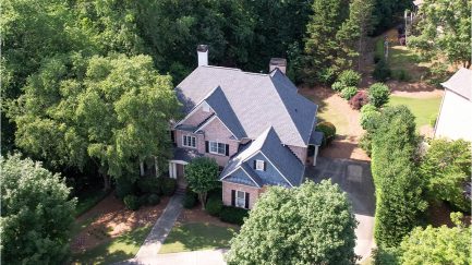 Aerial view of a two-story brick house with a gray roof, surrounded by trees and greenery, with a driveway leading to the entrance.
