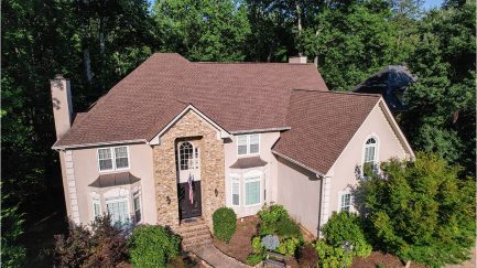 A two-story house with a steep brown shingle roof, stone and stucco exterior, arched windows, and a landscaped front yard, surrounded by trees.