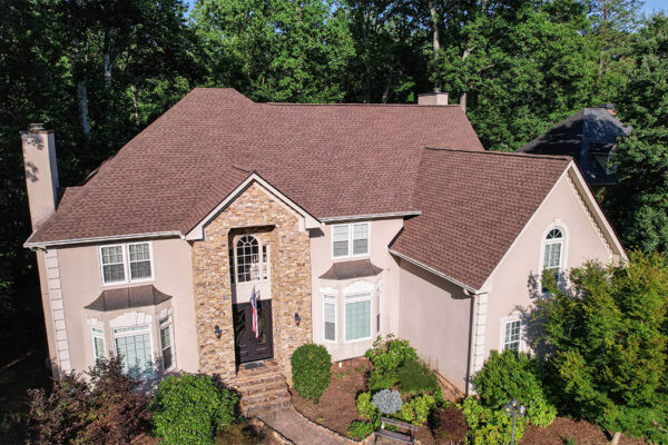 A two-story house with a steep brown shingle roof, stone and stucco exterior, arched windows, and a landscaped front yard, surrounded by trees. A two-story house with a steep brown shingle roof, stone and stucco exterior, arched windows, and a landscaped front yard, surrounded by trees.