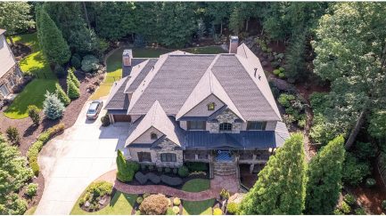 Aerial view of a large stone and brick house with a manicured lawn, curved driveway, landscaped garden, and surrounding trees.