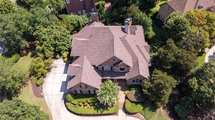 Aerial view of a large two-story brick house with a complex roof, surrounded by trees, lawn, and a curved driveway.