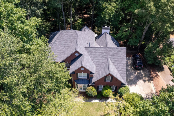 Aerial view of a two-story brick house with a gray roof, surrounded by trees, with a driveway and parked car to the right. Aerial view of a two-story brick house with a gray roof, surrounded by trees, with a driveway and parked car to the right.