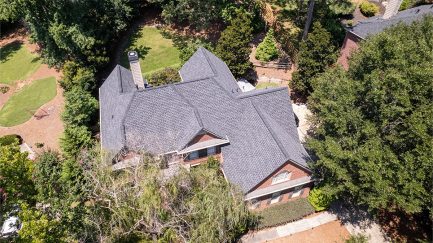 Aerial view of a large house with a dark gray shingle roof surrounded by trees and landscaped yard.