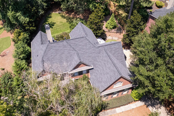 Aerial view of a large house with a dark gray shingle roof surrounded by trees and landscaped yard. Aerial view of a large house with a dark gray shingle roof surrounded by trees and landscaped yard.