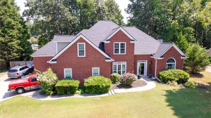 A two-story red brick house with multiple gables, surrounded by green shrubs and trees. A red pickup truck is parked in the driveway on the left.