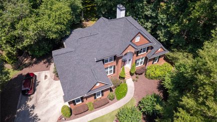 Aerial view of a two-story brick house with a dark shingled roof, surrounded by trees and landscaping, with a driveway and a red vehicle parked to the left.