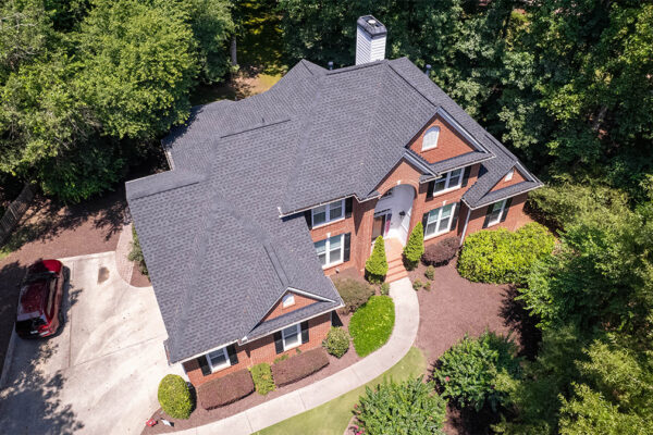 Aerial view of a two-story brick house with a dark shingled roof, surrounded by trees and landscaping, with a driveway and a red vehicle parked to the left. Aerial view of a two-story brick house with a dark shingled roof, surrounded by trees and landscaping, with a driveway and a red vehicle parked to the left.