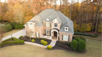 A large two-story brick house with gray shingles, arched windows, and landscaped bushes, set beside a driveway and surrounded by autumn trees.