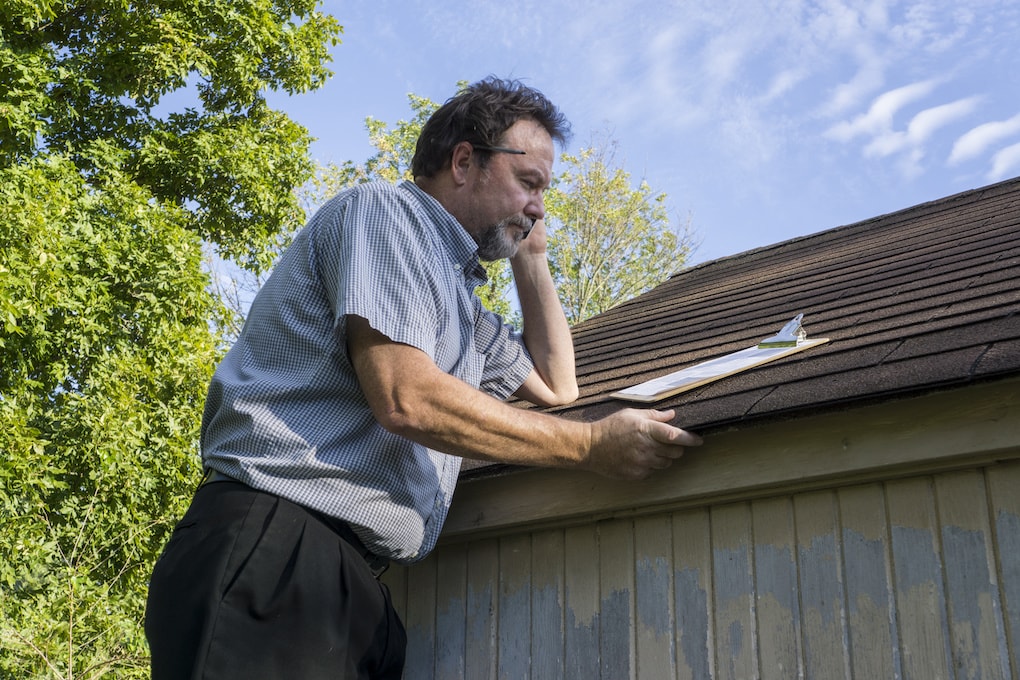  roof inspector holding clipboard inspecting storm-damaged shingle roof for insurance claim