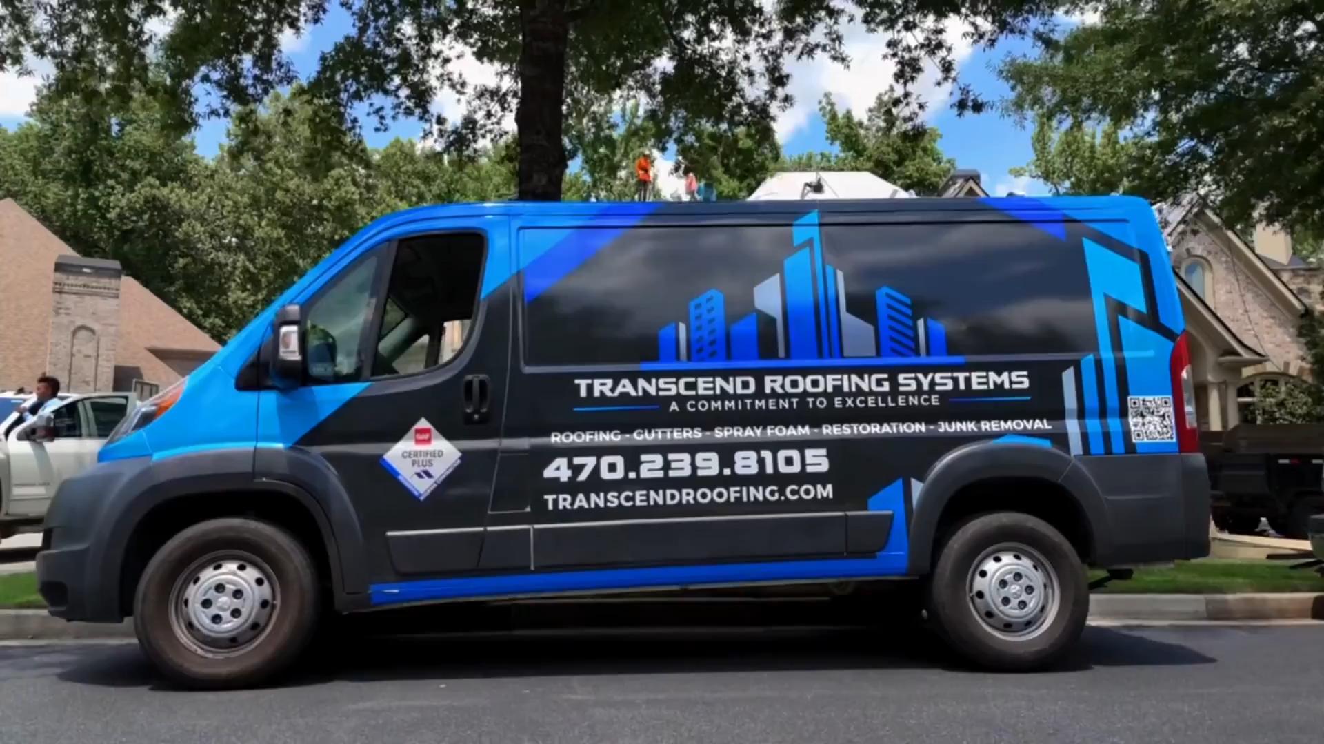 A blue and black van displaying the "Transcend Roofing Systems" logo, complete with contact info and building themed graphics, stands in the foreground  In the background, trees frame busy workers attending to their tasks 